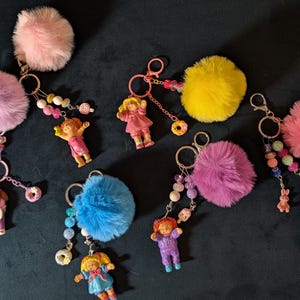 May include: A collection of keychains featuring colorful pom-poms in pink, purple, yellow, and blue. Each keychain has a small doll charm, some with beaded accents and miniature donut or candy charms. The keychains are on a dark background.