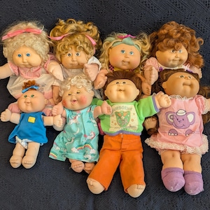 May include: A collection of eight Cabbage Patch Kids dolls. The dolls have various skin tones, hair colors, and outfits, including dresses, overalls, and pajamas. Some dolls have bows in their hair. The dolls are arranged on a dark blue surface.