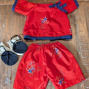 May include: A red short-sleeved top with dark blue trim and a side tie, matching red shorts with a floral pattern, and a pair of white socks with black circular felt soles. The fabric features a subtle dotted texture and blue floral accents.