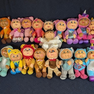 May include: A collection of seventeen Cabbage Patch Kids dolls arranged in two rows. Each doll features a soft fabric body, vinyl head with yarn hair, and is dressed in a unique animal-themed or patterned hooded outfit with various colors.