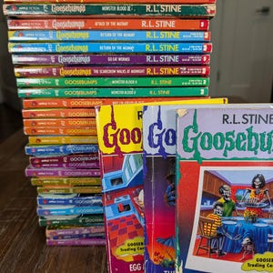 May include: A collection of Goosebumps books by R.L. Stine, featuring a tall stack of paperback spines with various colors and titles, alongside three upright books with illustrated covers showing skeletons and colorful graphic designs.