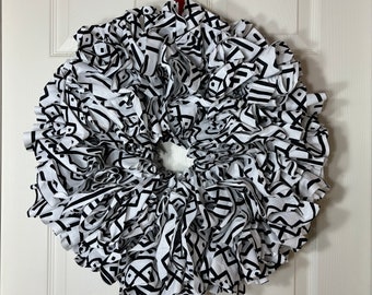 May include: A round wreath made of white fabric with a black geometric pattern. The wreath is hanging on a white door with a red ribbon. The wreath is a decorative item.