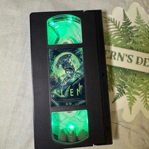 May include: A black VHS tape with a green light inside. The front of the tape has a picture of the alien from the movie "Alien" with the title "ALIEN" printed below it.