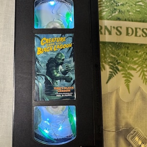 May include: A modified VHS tape featuring the film "Creature from the Black Lagoon." Fairy lights illuminate the top and bottom sections of the tape, with the movie cover displayed in the centre.