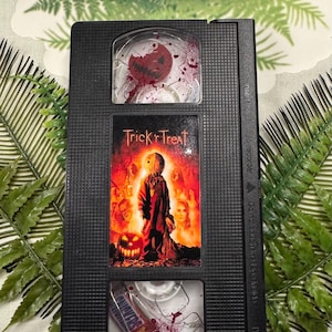 May include: Black VHS tape featuring the movie "Trick 'r Treat." The tape has a decorative design with a Halloween theme, including images of a jack-o'-lantern and a figure in a costume. The top of the tape has the text "FERN'S DESIGNS."