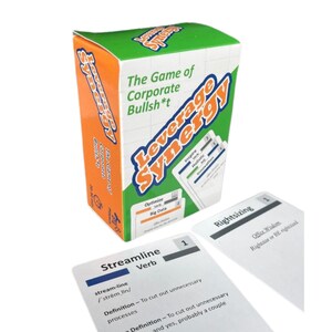 May include: A boxed game titled "Leverage Synergy" with orange and green packaging. The box reads "The Game of Corporate Bullsh*t." Several white cards with black text are displayed, including "Streamline" and "Rightsizing."