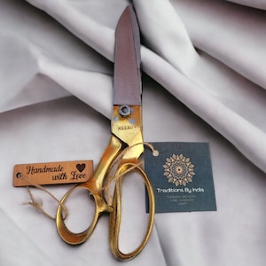 以下が含まれることがあります： 「Handmade with Love」と書かれた木製のタグと、「Traditions By India Timeless artistry time-honored craft」と書かれた黒いタグが付いた金色の布切りばさみ。