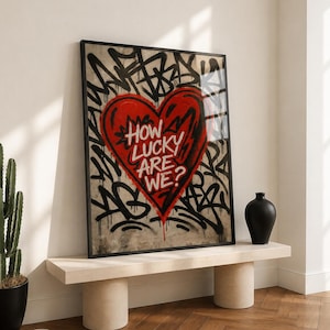 May include: A framed rectangular wall art piece featuring a large red heart centered on a beige background. The heart contains the white text "HOW LUCKY ARE WE?" surrounded by black graffiti-style markings and drips that extend across the entire print.