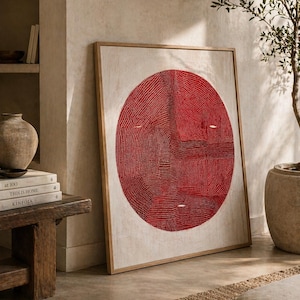 May include: Large framed artwork featuring a vibrant red circular design on a neutral background. The artwork is displayed in a light wooden frame, leaning against a wall. The room has a neutral color palette with natural elements.
