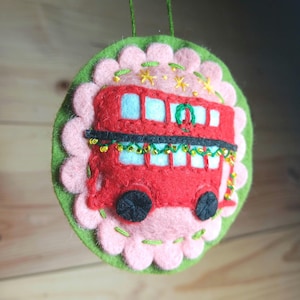 May include: A handmade felt Christmas ornament featuring a red double-decker bus. The bus has black wheels and blue windows, with a green garland. The ornament is surrounded by a pink scalloped border on a green felt base, with yellow star accents and a green hanging string.