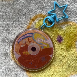 May include: A round, clear acrylic keychain with a cartoon illustration of a person with orange hair. The keychain has a blue star-shaped clasp and a blue ring. The text "FIRST WAVE" and Japanese text are visible on the keychain.
