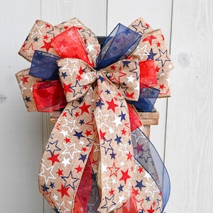 May include: A large decorative bow with burlap ribbon featuring red, white, and blue stars. The bow has multiple layers of ribbon in red, white, and blue, perfect for patriotic or holiday decor.