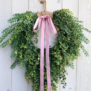 May include: A green leafy wreath with a pink ribbon hanging against a white wooden background. The wreath is made of faux greenery and has a natural, rustic look. The pink ribbon adds a touch of color and elegance to the wreath.