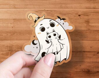Op de afbeelding: Een gestanste sticker met een vriendelijk spook met een heksenhoed, die een kat vasthoudt. Het spook en de kat zijn wit met zwarte details, tegen een bruine achtergrond met sterren, wolken en vleermuizen.