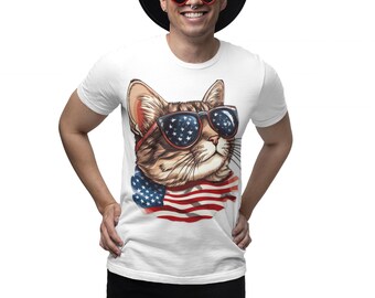 May include: A white t-shirt featuring a graphic print of a brown tabby cat wearing sunglasses with a star-spangled pattern. The cat is positioned above a draped American flag design. The shirt is paired with a black hat, red heart-shaped glasses, and black shorts.