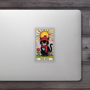 May include: A sticker featuring a black cat wearing a red hat and scarf, sitting in front of a yellow sun. The words "THE SUN" are at the bottom. The cat is surrounded by colorful flowers, creating a tarot card design.