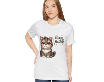 May include: White t-shirt featuring a cartoon kitten with a crown and the text "FEED ME, PEASANT!" in a speech bubble. The kitten is brown and gray with large eyes. The shirt is worn by a person.