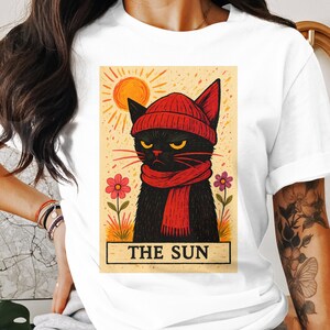 May include: White t-shirt featuring a graphic of a black cat wearing a red beanie and scarf, with a sun and flowers in the background. The text "THE SUN" is at the bottom of the image. The cat has yellow eyes and a grumpy expression.