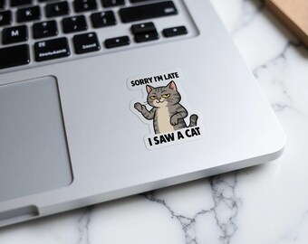 Può includere: Un adesivo con un gatto grigio tigrato che saluta con la zampa. Il testo dice "SORRY I'M LATE" e "I SAW A CAT". L'adesivo è posto su un computer portatile argentato.