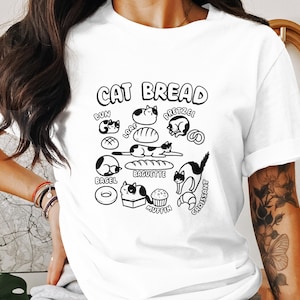 Puede incluir: Camiseta blanca con un diseño gráfico en blanco y negro con el texto "CAT BREAD" sobre ilustraciones de varios productos de panadería, cada uno con un gato incorporado en el diseño. Incluye un bagel, un cruasán y un pretzel.