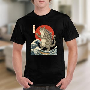 May include: Black t-shirt featuring a cat-monster design inspired by Japanese art. The illustration shows a cat with a Godzilla-like appearance standing on ocean waves in front of a red sun. The Japanese text is visible.