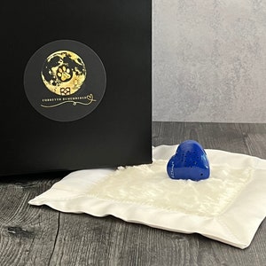 Könnte beinhalten: Ein weißes, flauschiges Herzförmiges Kissen mit einem blauen herzförmigen Soundgerät darauf. Das Soundgerät hat einen Pfotenabdruck und die Worte "Forever Remembered" darauf. Das Kissen liegt auf einer Holzoberfläche.