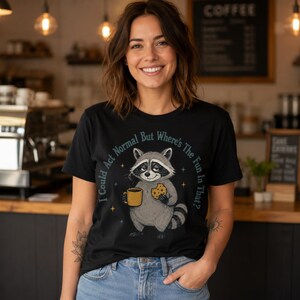 May include: A black t-shirt featuring a graphic of a raccoon holding a yellow coffee mug and a chocolate chip cookie. The text surrounding the raccoon reads, I Could Act Normal But Where's The Fun In That? The design includes small star accents.