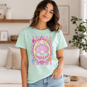 Peut inclure: Un t-shirt vert menthe clair avec un motif central représentant un coquillage à carreaux roses et blancs contenant une grosse perle blanche. Le dessin est encadré par une bordure festonnée, des nœuds roses et des étoiles colorées sur les côtés.