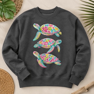 Peut inclure: Un sweat-shirt à col rond gris anthracite foncé orné d'une impression graphique représentant trois tortues de mer empilées verticalement. Chaque tortue présente un motif multicolore vif sur sa carapace, incluant des designs floraux, à carreaux et en chevrons.
