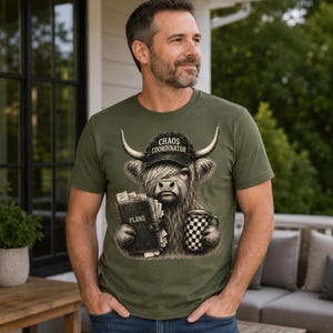 Peut inclure: Un t-shirt vert olive avec une illustration de vache Highland portant une casquette noire marquée "CHAOS COORDINATOR". La vache tient un agenda noir intitulé "PLANS" et une tasse à carreaux noirs et blancs dans ses sabots poilus.