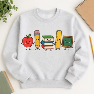 Peut inclure: Un sweat-shirt à col rond gris clair avec une impression graphique de cinq fournitures scolaires anthropomorphes se tenant par la main. Les personnages incluent une pomme rouge, un crayon jaune, trois livres empilés, une règle jaune et un tableau vert.