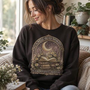Peut inclure: Un sweat-shirt à col rond marron foncé avec une impression graphique centrale représentant une pile de livres surmontée d'un livre ouvert, entourée de marguerites et de lavande sous un croissant de lune, avec le texte A WELL-READ WOMAN IS A DANGEROUS CREATURE.