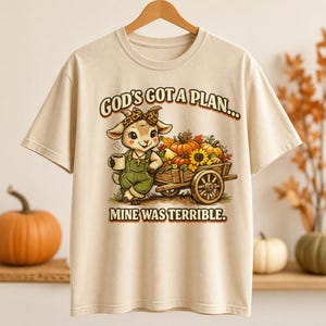 Peut inclure: Un t-shirt beige à manches courtes orné d'un dessin de chèvre portant un bandeau léopard et une salopette verte, tenant une tasse près d'une charrette en bois remplie de citrouilles et de tournesols, avec le texte GOD'S GOT A PLAN... MINE WAS TERRIBLE.