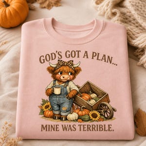 Peut inclure: Un sweat-shirt rose clair avec une illustration d'une vache highland duveteuse portant une salopette en jean et un nœud léopard, tenant une tasse. À côté de la vache se trouve une charrette en bois remplie de citrouilles. Le texte indique GOD'S GOT A PLAN... MINE WAS TERRIBLE.