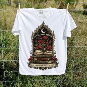 Peut inclure: Un t-shirt blanc à manches courtes avec une impression graphique représentant un livre ouvert d'où poussent des roses rouges, encadré par une arche gothique ornée. Le texte indique A WELL-READ WOMAN IS A DANGEROUS CREATURE, avec un croissant de lune.