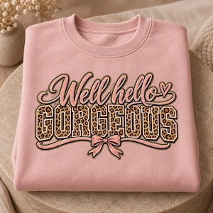 Peut inclure: Un sweat-shirt à col rond rose clair avec le texte Well hello GORGEOUS écrit en lettres cursives et en lettres capitales. Le mot GORGEOUS est rempli d'un motif léopard marron et noir. Un petit cœur rose et un nœud rose complètent le motif.