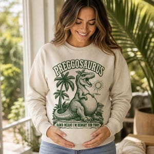 Peut inclure: Un sweat-shirt beige à col rond avec un motif vert foncé représentant un dinosaure portant un bandeau. Le texte indique PREGGOSAURUS et RAWR MEANS I'M HUNGRY FOR TWO. L'illustration comprend des palmiers et un soleil, centré sur la poitrine.