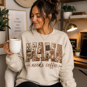 Peut inclure: Un sweat-shirt beige à col rond arborant le mot MAMA en grandes lettres majuscules remplies de motifs sur le thème du café, incluant des grains, des tasses et des fleurs, avec le texte needs coffee écrit en cursive sous les lettres.