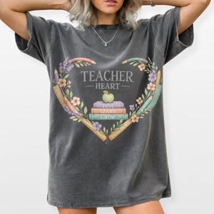 Peut inclure: Un t-shirt gris anthracite à manches courtes orné d'un motif en forme de cœur composé de crayons, de règles et de fleurs. Au centre, trois livres empilés marqués LOVE, LEARN, GROW sont surmontés d'une pomme verte et du texte TEACHER HEART.