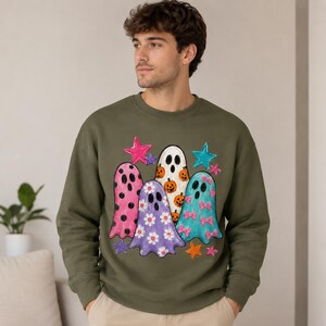 Peut inclure: Un sweat-shirt à col rond vert olive orné d'un motif imprimé représentant quatre fantômes. Les fantômes sont décorés de pois roses, de marguerites blanches, de citrouilles orange et de nœuds roses. Le motif inclut des étoiles colorées.