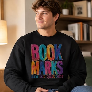 Peut inclure: Un sweat-shirt noir à col rond avec une impression graphique colorée sur la poitrine. Le texte indique BOOK MARKS en grandes lettres texturées multicolores, avec la phrase are for quitters en plus petites lettres multicolores en dessous.