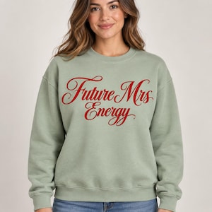 Peut inclure: Un sweat-shirt à col rond vert sauge avec le texte Future Mrs Energy imprimé en lettres cursives rouges audacieuses sur la poitrine. Le vêtement présente des manches longues, un col côtelé et des poignets côtelés, offrant une coupe standard.