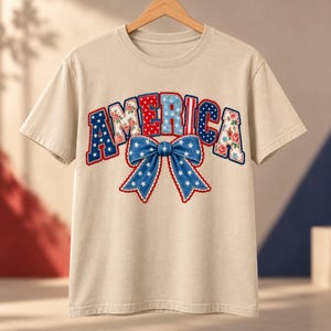 Peut inclure: Un t-shirt beige à manches courtes arborant le mot AMERICA dans une police à motifs variés aux couleurs rouge, blanc et bleu, incluant des designs floraux et à pois. Sous le texte se trouve un grand nœud bleu avec des étoiles blanches.