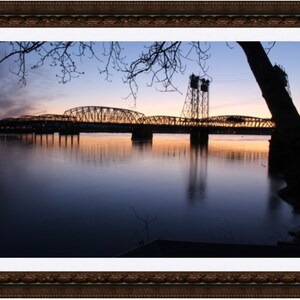 May include: A framed landscape photograph of a bridge over a calm body of water at dusk. The sky transitions from blue to orange, reflecting in the water. Silhouetted trees and bridge structures add depth.