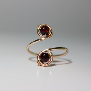 May include: A gold ring with two red garnet stones set in wire wrapped settings. The ring is a simple, minimalist design.