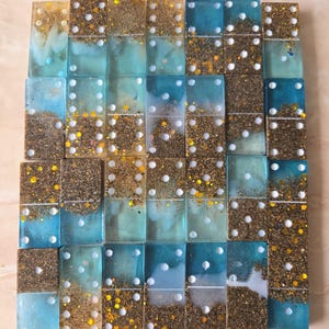 May include: A set of rectangular dominoes made of translucent resin in shades of blue and clear, featuring embedded gold glitter and sand-like particles. Each piece displays white circular pips arranged in standard numerical patterns across the surface.