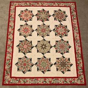 May include: A rectangular quilt featuring a grid of twelve star-shaped blocks on a cream background. Each star displays intricate floral and botanical patterns in shades of red, green, and brown. The quilt is framed by a red border and a floral edge.