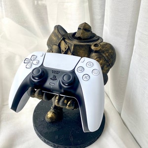 Könnte beinhalten: Eine goldfarbene, 3D-gedruckte Figur eines Roboters, der einen weißen PlayStation 5-Controller hält. Der Roboter steht auf einem schwarzen runden Sockel mit einer strukturierten Oberfläche.