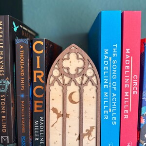 Könnte beinhalten: Ein hölzerner Buchstütze mit einem gotischen Fensterdesign, verziert mit Mond, Sternen und Drachensilhouetten. Sie steht zwischen Büchern wie "Circe" und "The Song of Achilles" von Madeline Miller. Die Bücher haben blaue, rote und schwarze Buchrücken.