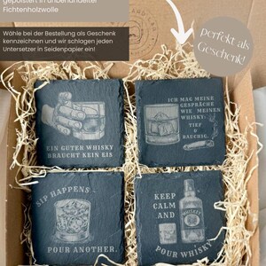 May include: A set of four square slate coasters with engraved whiskey-themed designs and phrases. The coasters are packaged in a cardboard box with decorative straw. Phrases include "Ein Guter Whisky Braucht Kein Eis" and "Keep Calm and Pour Whisky."